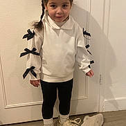 Elya participe au concours pour gagner de l'argent avec cette photo : black_bows, black_leggings, braided_hair, casual_clothing, child, cozy, cute, door, fashion, floor, girl, indoor, plush_hat, portrait, smiling, sneakers, standing, white_sweater, wooden_floor, young_child