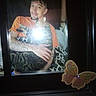 bed, blanket, butterfly_sticker, cat, dark_background, flash_glare, hand, holding, home_decor, indoor, man, on_chest, orange_shirt, person, picture_frame, pillow, portrait, smiling, tabby_cat, tattoos