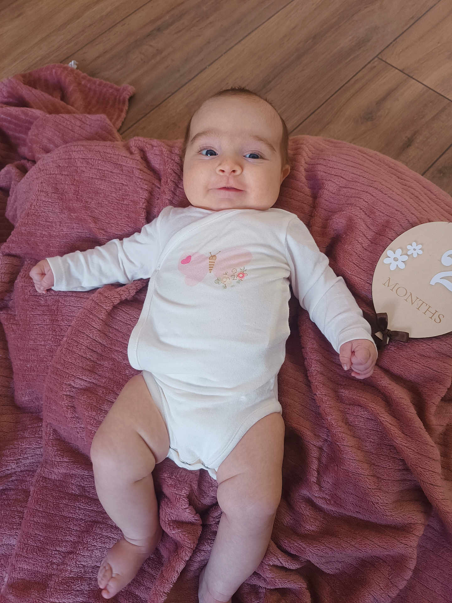 Lila a rejoint le concours — aidez-le/la à gagner de superbes lots ! baby, infant, blanket, floor, wooden_floor, onesie, butterfly, plaque, milestone, pink, white_clothing, cute, lying_down, chubby, young_child, smiling, indoors, soft_texture, milestone_marker, portrait