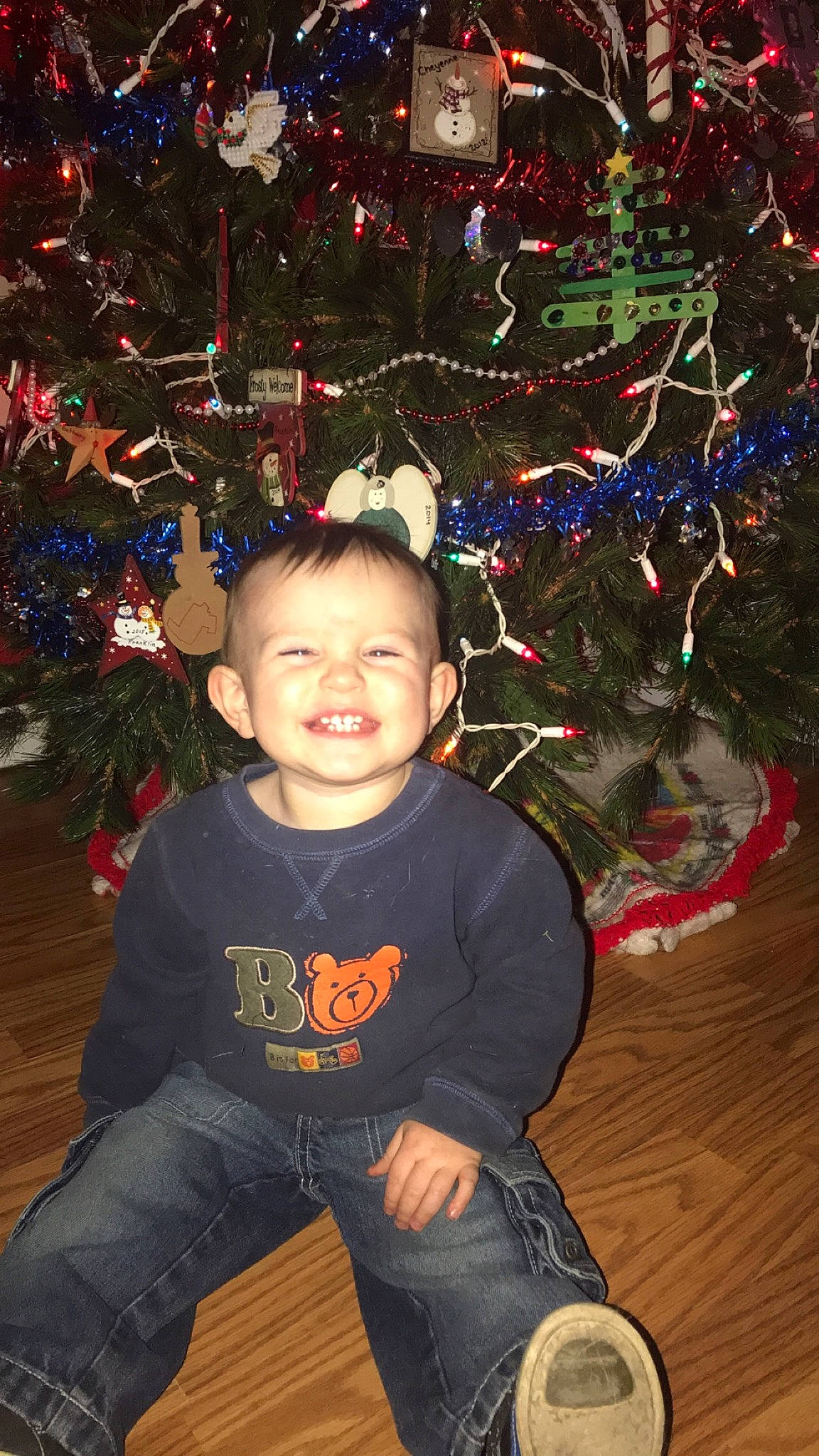 Kota is registered to the contest to win money with this photo: child, christmas, christmas_decoration, christmas_eve, christmas_lights, christmas_ornament, christmas_tree, event, fir, happy, holiday, holiday_ornament, interior_design, joy, person, plant, sitting, smile, toddler, tree