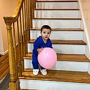Muhammad is registered to the contest to win money with this photo: toddler, balloon, stairs, wooden_stairs, blue_outfit, slippers, indoor, handrail, wooden_floor, child, curious, pink_balloon, home_interior, person, smiling, standing, railing, footwear, young_child, face