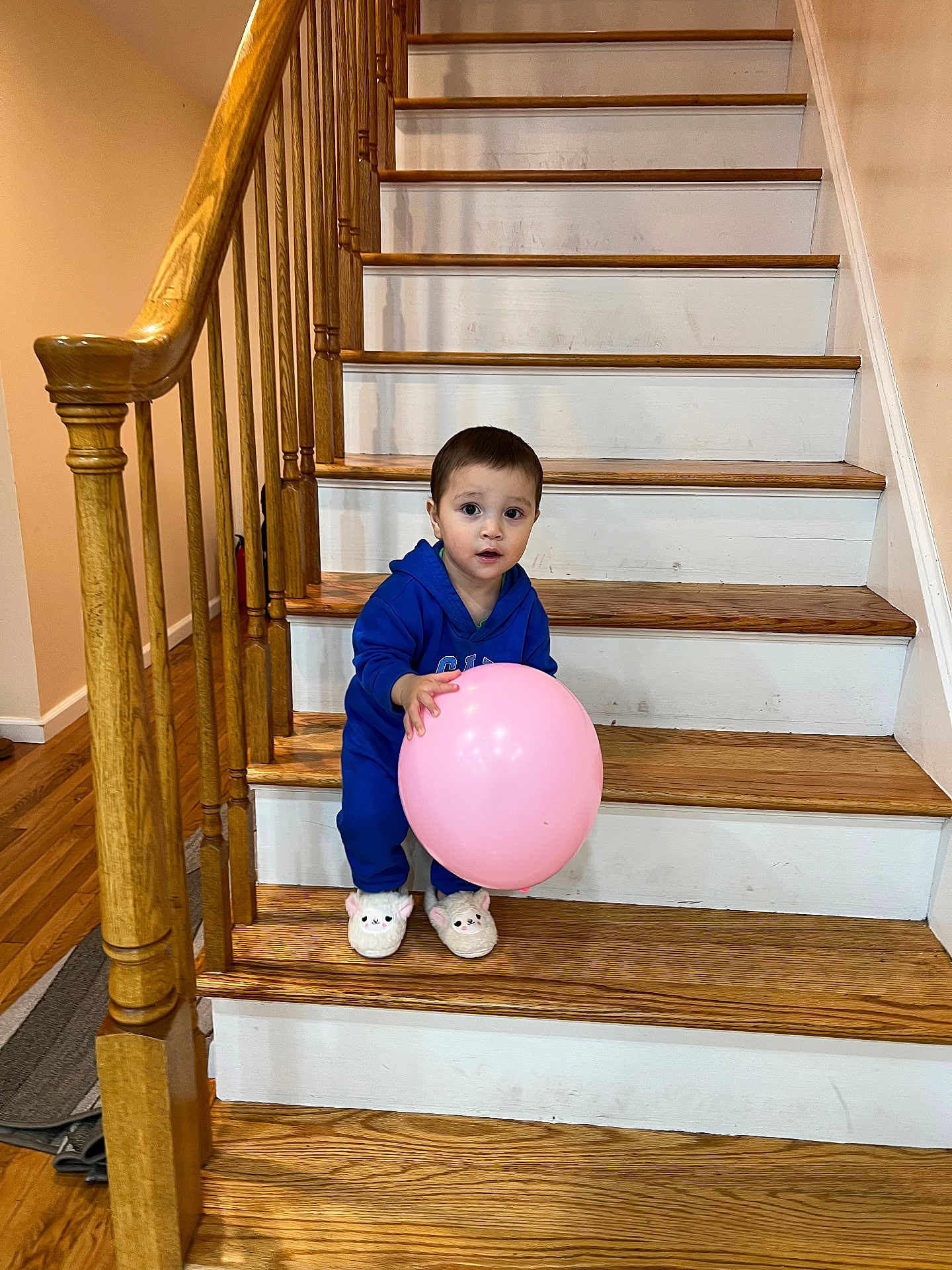 Muhammad is registered to the contest to win money with this photo: toddler, balloon, stairs, wooden_stairs, blue_outfit, slippers, indoor, handrail, wooden_floor, child, curious, pink_balloon, home_interior, person, smiling, standing, railing, footwear, young_child, face