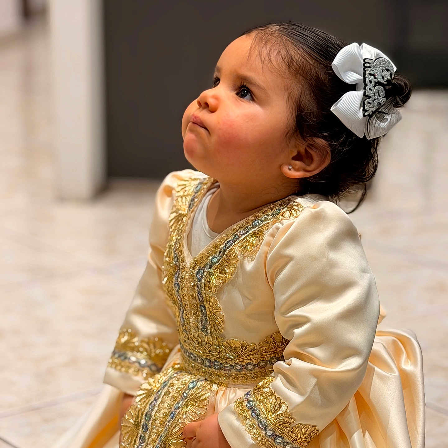 Eyana participe au concours pour gagner de l'argent avec cette photo : bow, candid, child, cute, dress, earrings, formal_dress, girl, gold_embroidered, hair_bow, indoor, looking_up, ornate, portrait, pout, profile, sitting, soft_background, tile_floor, toddler