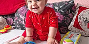 Sofia is registered to the contest to win money with this photo: baby, child, red_onesie, toy, stuffed_animal, ball, bed, pillow, floral_pattern, smile, happy, indoors, cute, infant, person, playful, toddler, hair_ponytail, sitting, home
