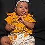 baby, child, tongue_out, yellow_shirt, floral_shorts, headband, white_bow, sitting, couch, sandals, playful, smiling, indoor, person, cute, infant, clothing, footwear, happy, portrait