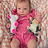 baby, infant, pink_outfit, plush_toy, unicorn, cow_toy, blanket, checkered, cute, child, indoors, soft_texture, smiling, face, hand, legs, comfort, resting, colorful, portrait