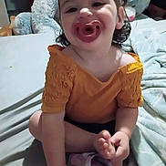 Elizabeth joined the competition — help win amazing prizes! toddler, child, pacifier, orange_clothing, bed, plush_toy, teddy_bear, shoe, foot, hand, indoor, smiling, happy, person, curly_hair, blanket, furniture, cute, playful, cozy
