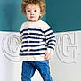 toddler, child, curly_hair, striped_sweater, jeans, barefoot, green_cushion, indoor, standing, curious, light_skin, blue_wall, white_wall, casual_clothing, portrait, young_child, cute, home, cushion, fashion