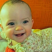 Scarlett is registered to the contest to win money with this photo: baby, smiling, face, infant, floral_outfit, lying_down, tummy, happy, cute, child, indoors, bright, orange_background, skin, person, young_child, closeup, portrait, cheerful, expression