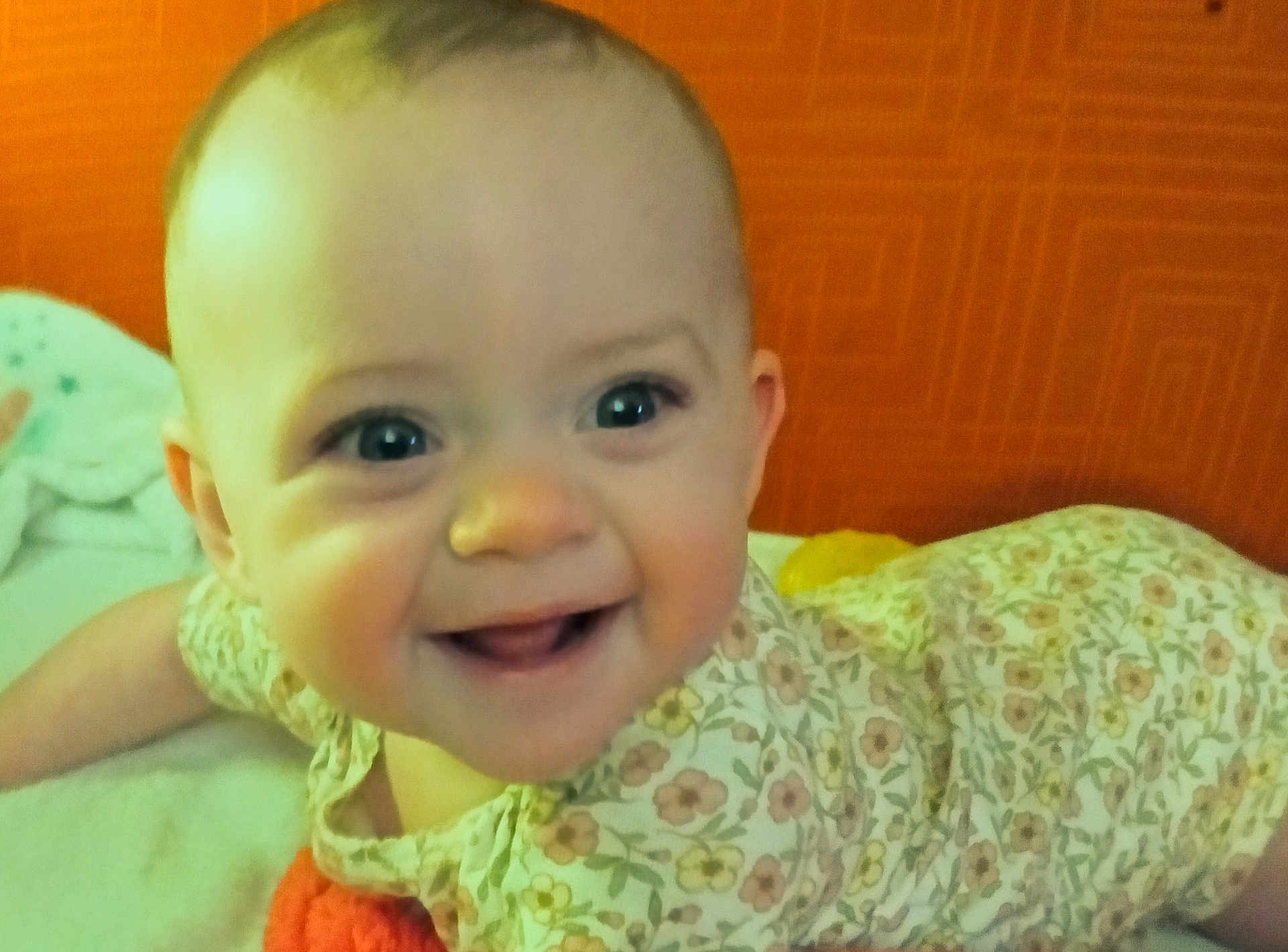 Scarlett is registered to the contest to win money with this photo: baby, smiling, face, infant, floral_outfit, lying_down, tummy, happy, cute, child, indoors, bright, orange_background, skin, person, young_child, closeup, portrait, cheerful, expression
