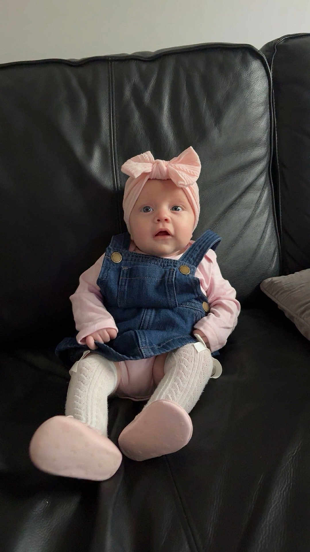 Amelia joined the competition — help win amazing prizes! baby, child, denim_dress, pink_headwrap, white_tights, pink_shoes, black_leather_couch, infant, sitting, cute, portrait, indoors, expression, small_hands, footwear, clothing, face, person, young_child, fashion
