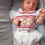 Tommy joined the competition — help win amazing prizes! baby, infant, smile, white_clothing, red_embroidery, cushion, textured_fabric, lying_down, portrait, cute, child, small_hands, pale_skin, indoor, soft_lighting, comfortable, happy, face, head, young