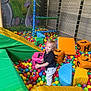 toddler, child, ball_pit, foam_blocks, colorful_balls, play_area, netting, indoor_playground, happy, smiling, black_shirt, white_pants, yellow, green, blue, orange, pink, purple, red, play