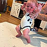 Braley is registered to the contest to win money with this photo: baby, infant, child, portrait, standing, supported, hands_raised, smiling, indoors, living_room, play_mat, star_pattern, onesie, diaper, barefoot, adult, tattoos, sneakers, television, furniture
