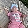 baby, infant, child, portrait, lying_down, outdoor, blanket, grass, pink_clothing, white_headband, bow, baby_girl, face, hands, arms, legs, text_on_clothing, fashion, natural_light, closeup