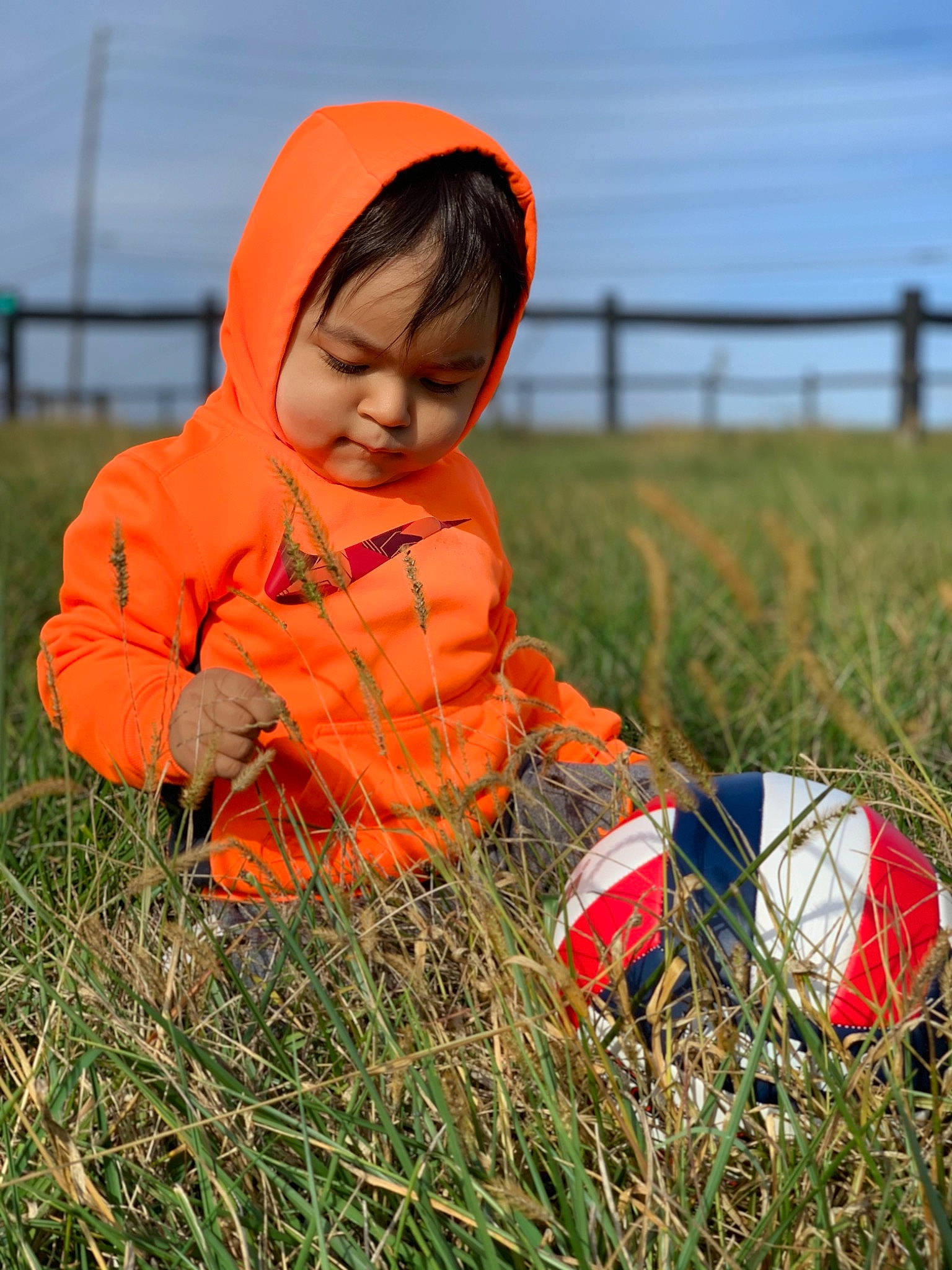 Mahir joined the competition — help win amazing prizes! baby, baby_toddler_clothing, ball, child, farm, field, football, grass, grassland, hood, hoodie, human, nose, orange, people_in_nature, person, play, prairie, soccer_ball, toddler