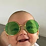 accessory, baby, bib, cheeks, close_up, clothing, eyes, eyewear, face, green_sunglasses, grin, happy, head, indoor, infant, person, portrait, smile, sunglasses, wall