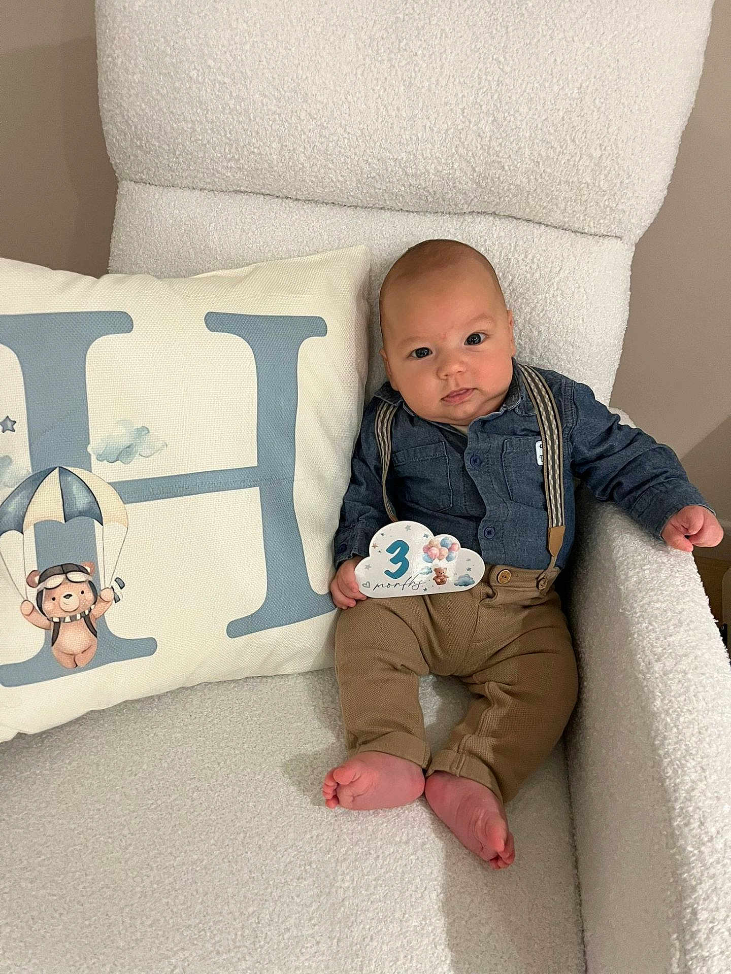Henri a rejoint le concours — aidez-le/la à gagner de superbes lots ! baby, infant, milestone_card, 3_months, denim_shirt, suspenders, brown_pants, barefoot, white_chair, fluffy_texture, decorative_pillow, letter_h, parachuting_bear, cute, portrait, indoors, smiling, child, relaxed, cozy