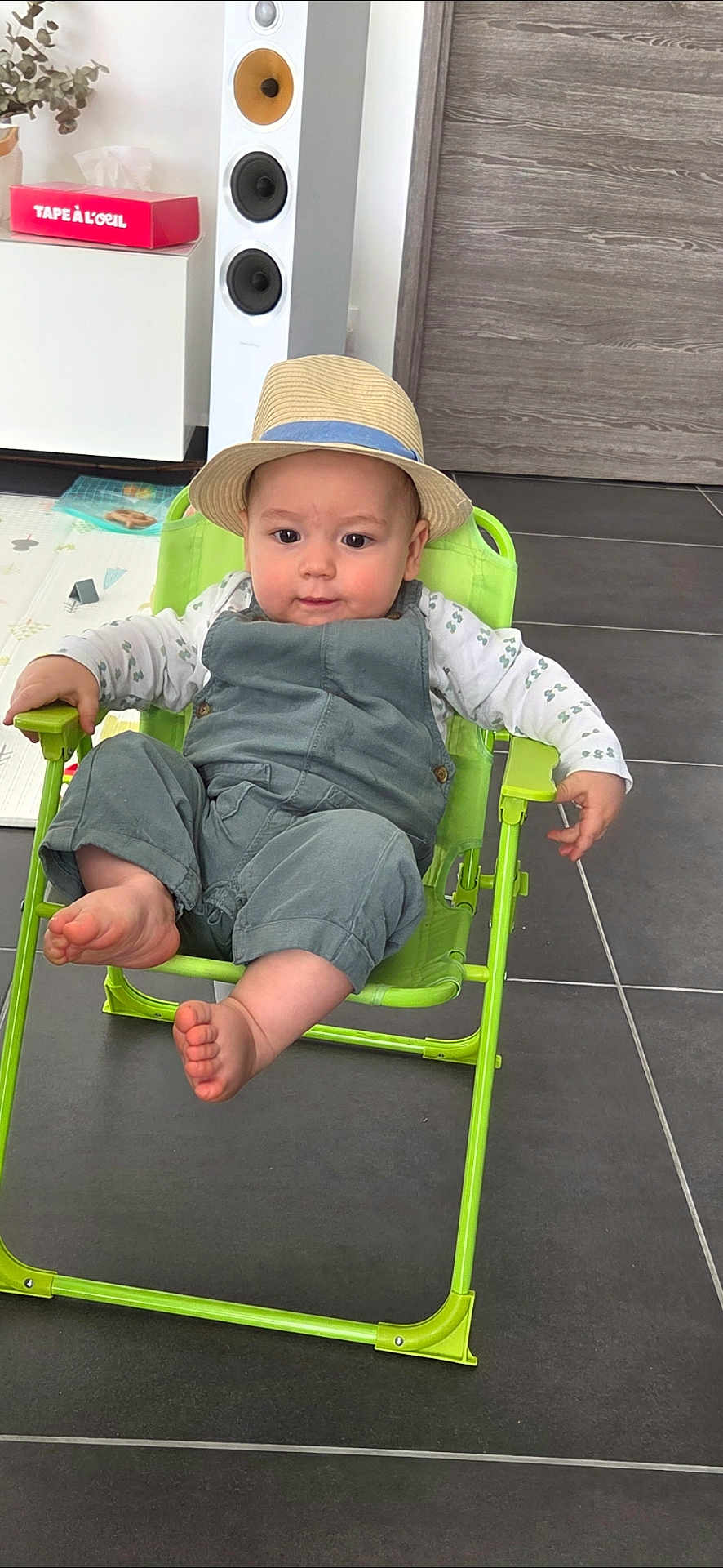 Henri participe au concours pour gagner de l'argent avec cette photo : baby, barefoot, cabinet, clothing, cute, face, feet, green_chair, hat, indoor, infant, overalls, playmat, portrait, smile, speaker, straw_hat, tiled_floor, toy, wooden_panel