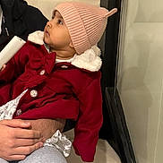 Fatimah is registered to the contest to win money with this photo: toddler, child, baby, hat, coat, red_clothing, pink_hat, indoor, person, hand, face, curious, warm_clothing, holding_object, adult, lap, seated, portrait, casual, cozy