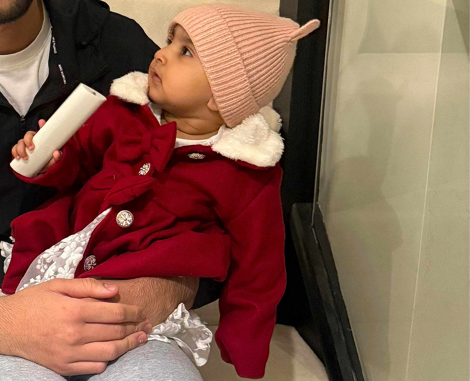 Fatimah is registered to the contest to win money with this photo: toddler, child, baby, hat, coat, red_clothing, pink_hat, indoor, person, hand, face, curious, warm_clothing, holding_object, adult, lap, seated, portrait, casual, cozy