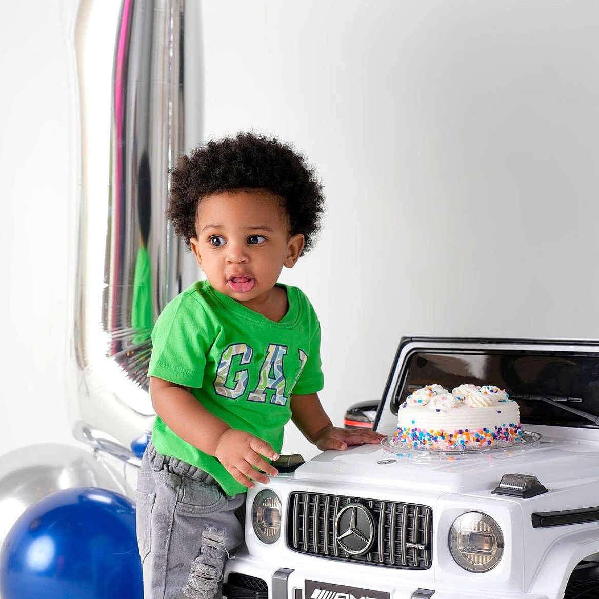 Nairobi is registered to the contest to win money with this photo: child, toddler, birthday, cake, balloons, toy_car, luxury_car, mercedes, amg, green_shirt, jeans, curly_hair, standing, indoors, celebration, sprinkles, blue_balloon, silver_balloon, portrait, playroom