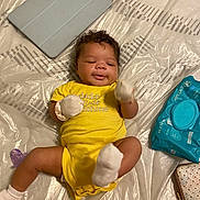 Alani joined the competition — help win amazing prizes! baby, infant, yellow_onesie, mittens, socks, pacifier, tablet, baby_wipes, plastic_sheet, lying_down, smiling, curly_hair, indoor, floor, cute, child, playful, small, person, cozy