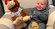 Roman is registered to the contest to win money with this photo: armchair, baby, baseball_cap, bedding, child, cozy, crib, cute, floral_pattern, happy, indoor, infant, monkey, plush_toy, portrait, seated, sign, smiling, striped_pajamas, three_months