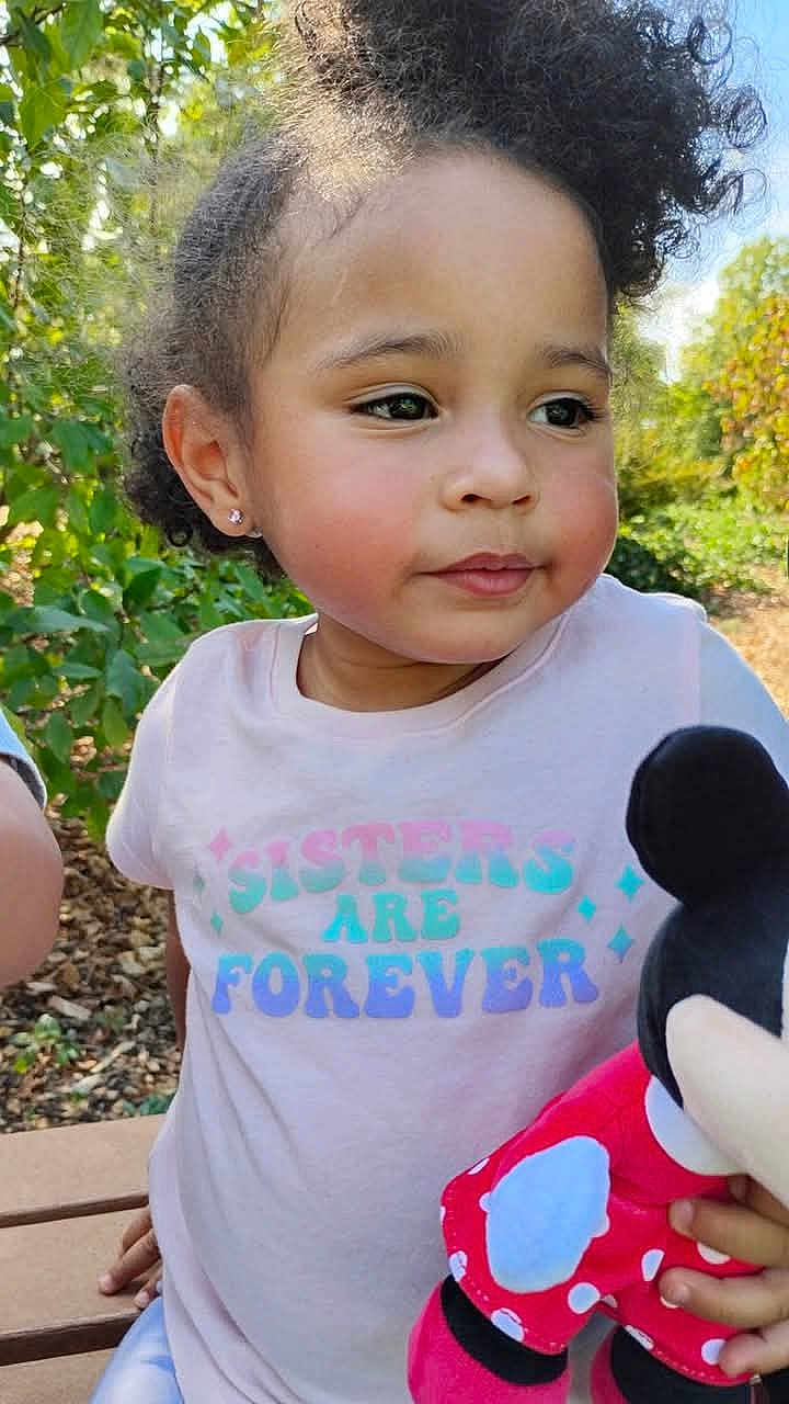 Amaya is registered to the contest to win money with this photo: child, girl, curly_hair, outdoor, bench, tshirt, plush_toy, mickey_mouse, greenery, sunlight, rosy_cheeks, portrait, casual_clothing, smiling, earrings, nature, daylight, cute, sitting, playful