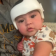 Adrian is registered to the contest to win money with this photo: baby, infant, child, helmet, protective_gear, onesie, animal_print, person, face, eyes, cheeks, hand, shirt, gray_clothing, indoor, couch, upclose, curious, portrait, holding
