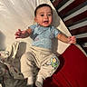 Adrian is registered to the contest to win money with this photo: baby, crib, blanket, blue_shirt, beige_pants, buzz_lightyear_patch, smiling, happy, reaching_out, socks, wooden_crib, cushion, infant, child, indoors, portrait, cute, clothing, person, face