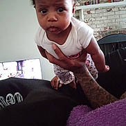 Neesa is registered to the contest to win money with this photo: baby, infant, child, person, tattoo, hand, white_shirt, patterned_pants, indoor, living_room, brick_wall, fireplace, television, furniture, curious, wide_eyes, holding, purple_blanket, casual_clothing, home
