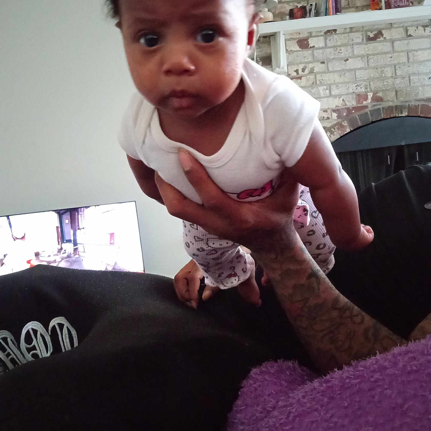 Neesa is registered to the contest to win money with this photo: baby, infant, child, person, tattoo, hand, white_shirt, patterned_pants, indoor, living_room, brick_wall, fireplace, television, furniture, curious, wide_eyes, holding, purple_blanket, casual_clothing, home
