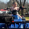 Leila joined the competition — help win amazing prizes! child, atv, blue_vehicle, outdoor, tree, bag, handlebars, playful, curious, vehicle, parked_car, nature, daylight, person, young, ponytail, grip, front_view, background_blur, adventure