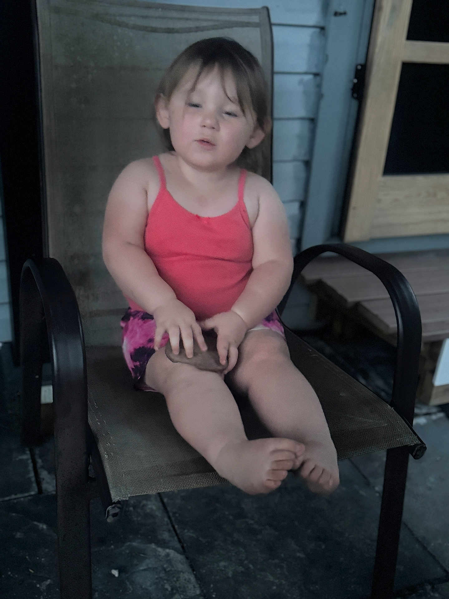 Leila is registered to the contest to win money with this photo: toddler, child, pink_top, shorts, chair, outdoor, porch, stone_floor, wooden_door, sitting, barefoot, face, hands, legs, expression, object, mesh_chair, casual, daylight, person