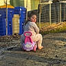 Leila is registered to the contest to win money with this photo: child, toddler, pajamas, outdoor, ride_on_toy, pink, purple, toy, face, looking_back, dirt_path, fence, bins, yellow, blue, curious, playful, daylight, person, casual