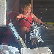 Leila joined the competition — help win amazing prizes! child, toddler, dirt_bike, motorcycle, red_tank_top, garage, clutter, boxes, tools, concentration, indoor, toy_vehicle, number_11, small_bike, person, young_child, grip, seat, handlebars, casual_clothing