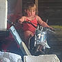 child, toddler, dirt_bike, motorcycle, red_tank_top, garage, clutter, boxes, tools, concentration, indoor, toy_vehicle, number_11, small_bike, person, young_child, grip, seat, handlebars, casual_clothing