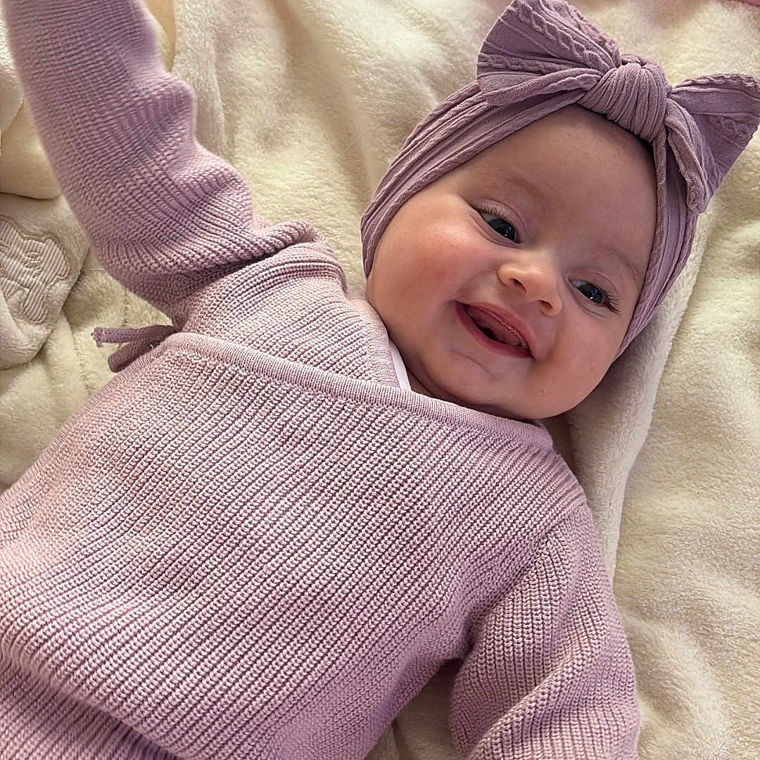 Lyana participe au concours pour gagner de l'argent avec cette photo : baby, blanket, bow, child, cozy, cute, face, happy, headband, indoor, infant, knitted_clothing, laying_down, person, pink, portrait, purple, smile, soft_texture, warm_clothing