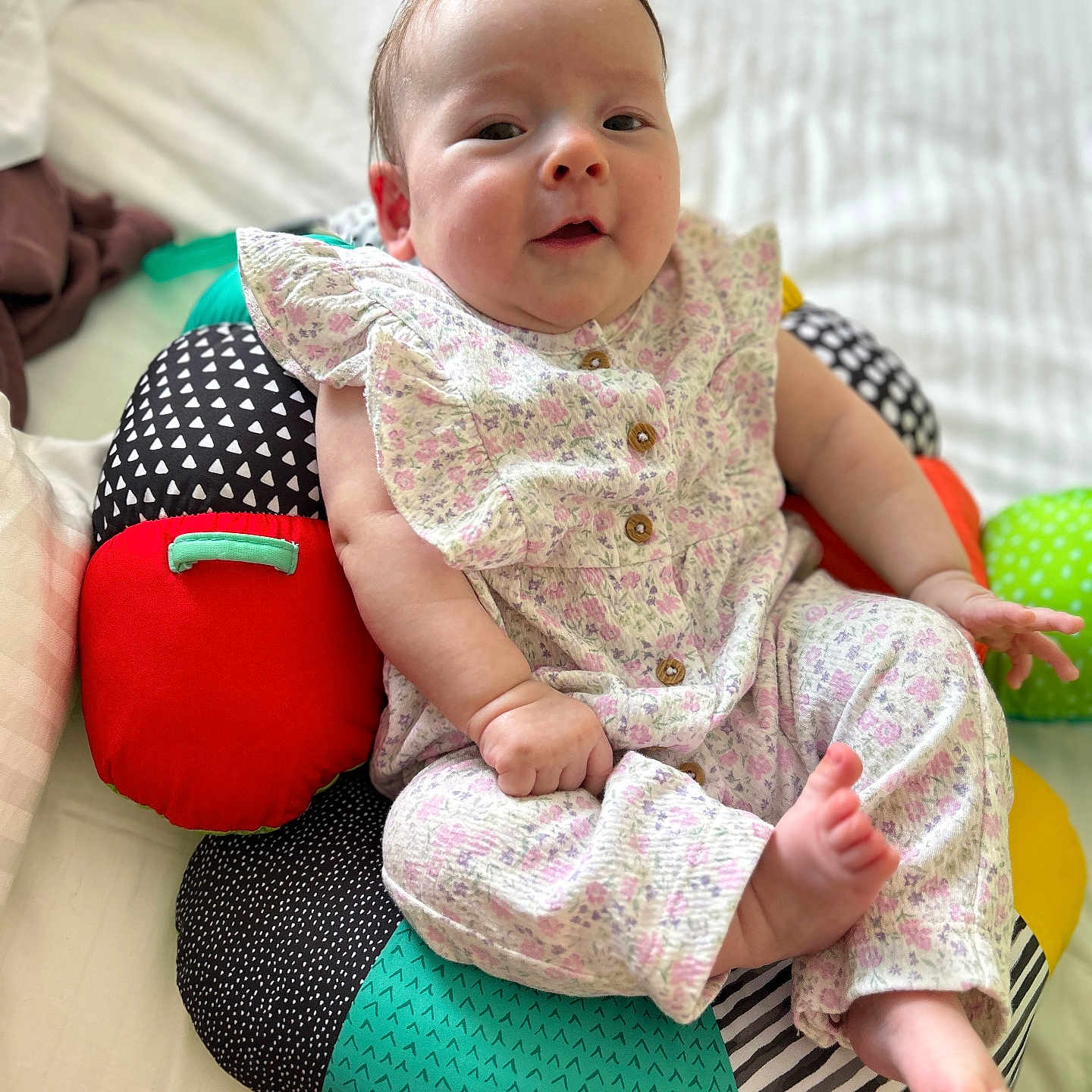 Isabella Larimore is registered to the contest to win money with this photo: baby, barefoot, bed, child, colorful, cushion, cute, face, floral_outfit, head, indoors, infant, patterned, person, plush, relaxed, small_hand, smile, white_bedspread, wooden_buttons