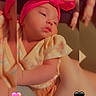 baby, headband, pink, yellow_outfit, floral_pattern, infant, sleepy, closeup, hand, arm, skin, cute, heart_emoji, blurred_background, portrait, indoors, soft_lighting, child, candid, love