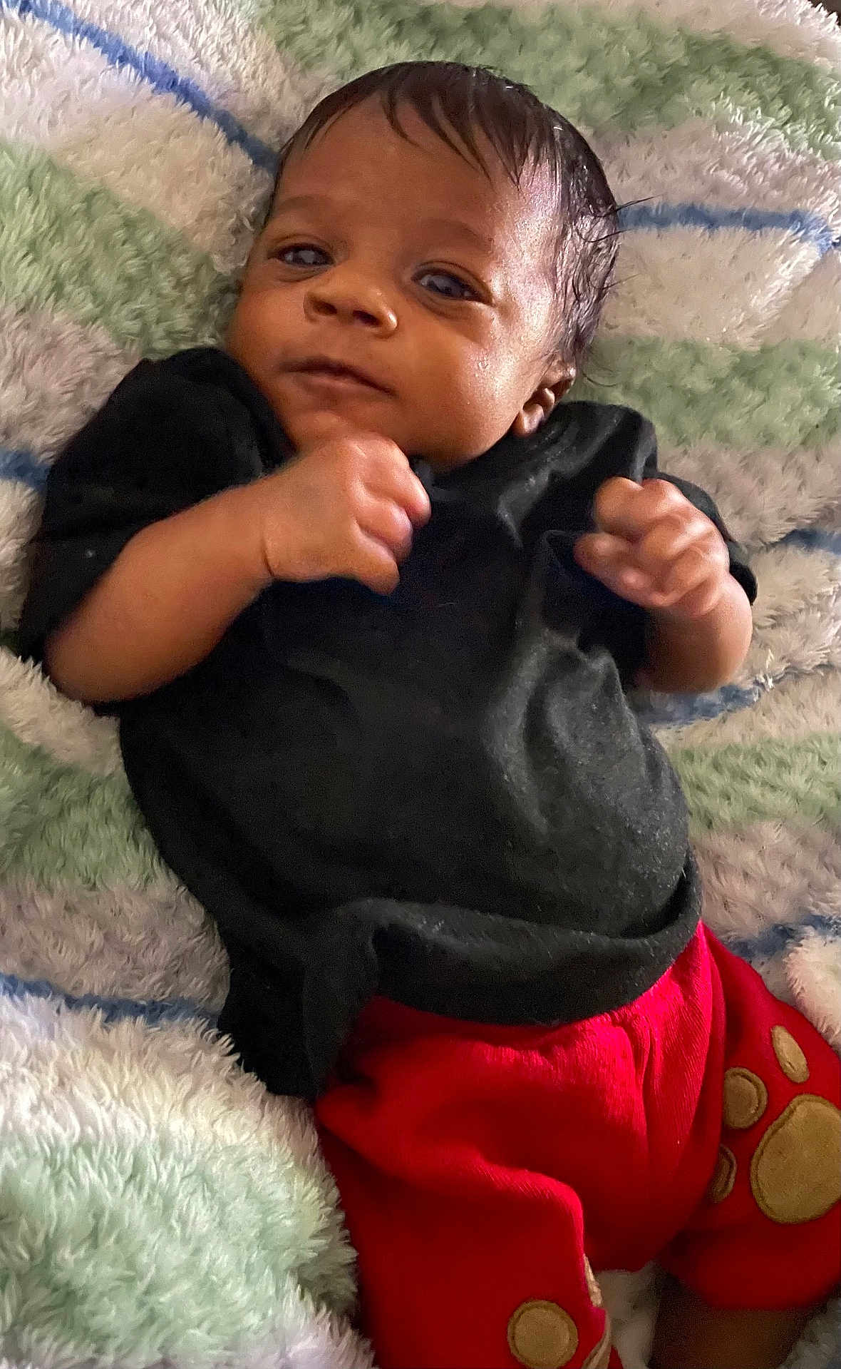 Josiah is registered to the contest to win money with this photo: baby, infant, child, black_shirt, red_pants, blanket, striped_blanket, curly_hair, fist, lying_down, cute, portrait, indoors, soft_texture, young_child, small_hands, expression, face, person, cozy