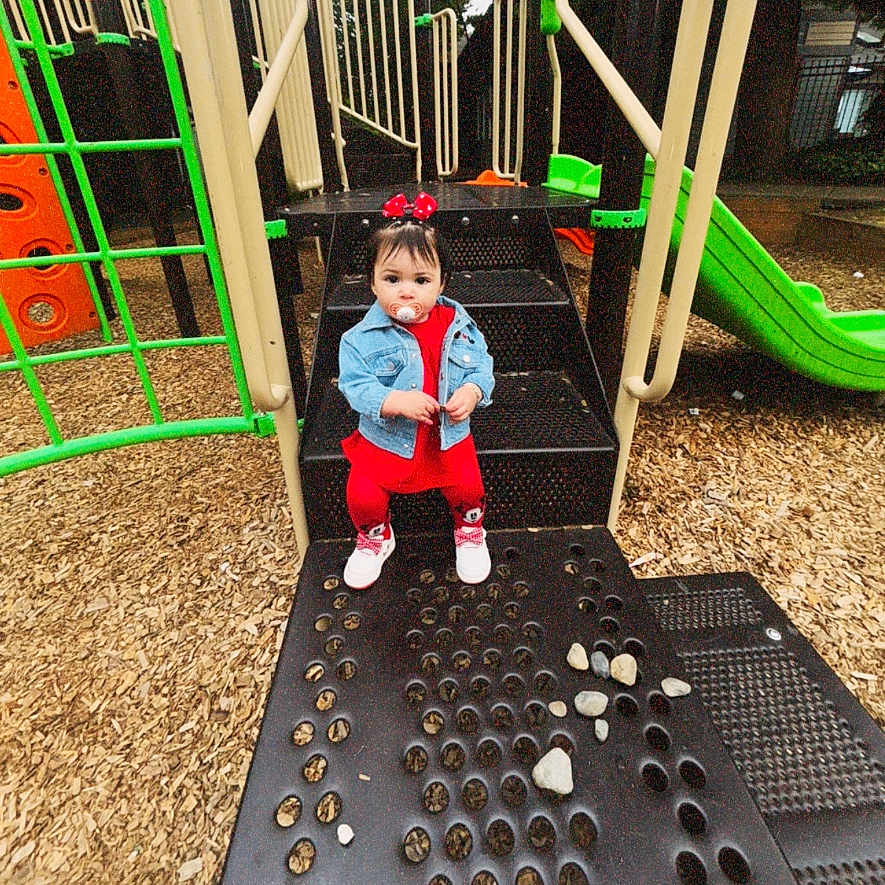 Lia Isabella is registered to the contest to win money with this photo: baby_shoes, black, bow, child, curious, daylight, denim_jacket, green, orange, outdoor, pacifier, person, playground, playground_equipment, red_clothing, sitting, slide, steps, toddler, woodchips
