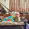 Dakota joined the competition — help win amazing prizes! baby, child, playpen, toy, indoor, curtains, couch, fishing_rods, plaid_shirt, person, furniture, blanket, living_room, face, infant, expression, seated, colorful, looking_away, home