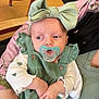 baby, infant, pacifier, headband, bow, green_dress, corduroy, button, hands, adult_hand, car_seat, blanket, pink_pattern, wooden_floor, table, chair_leg, jacket, portrait, side_glance, indoor