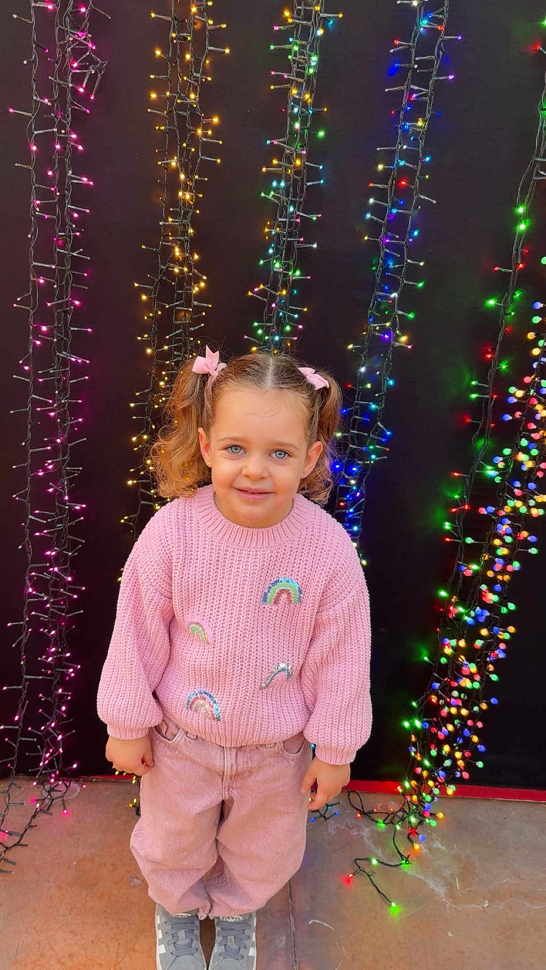Kessy participe au concours pour gagner de l'argent avec cette photo : child, girl, smiling, pink_sweater, rainbow, curly_hair, pigtails, fairy_lights, colorful_lights, background, outdoor, casual_clothing, pants, sneakers, happy, portrait, standing, cute, blue_eyes, festive