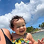 toddler, child, smiling, curly_hair, swimsuit, colorful_clothing, beach, water, ocean, sky, clouds, sunny, outdoor, person, holding, vacation, summer, nature, happy, portrait