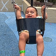 Shivansh is registered to the contest to win money with this photo: baby, black_swing, blue_surface, casual_clothing, child, feet, fun, green_wristband, happy, holding, infant, legs, metal_chains, outdoor, plants, playground, smiling, sunlight, swing, wall