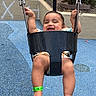 baby, black_swing, blue_surface, casual_clothing, child, feet, fun, green_wristband, happy, holding, infant, legs, metal_chains, outdoor, plants, playground, smiling, sunlight, swing, wall