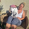 dog, woman, pet, outdoor, patio, wicker_chair, pink_hat, white_dog, sitting, lap, portrait, sunlight, shadows, wall, summer, casual_clothing, gray_pants, home, companion, funny_hat